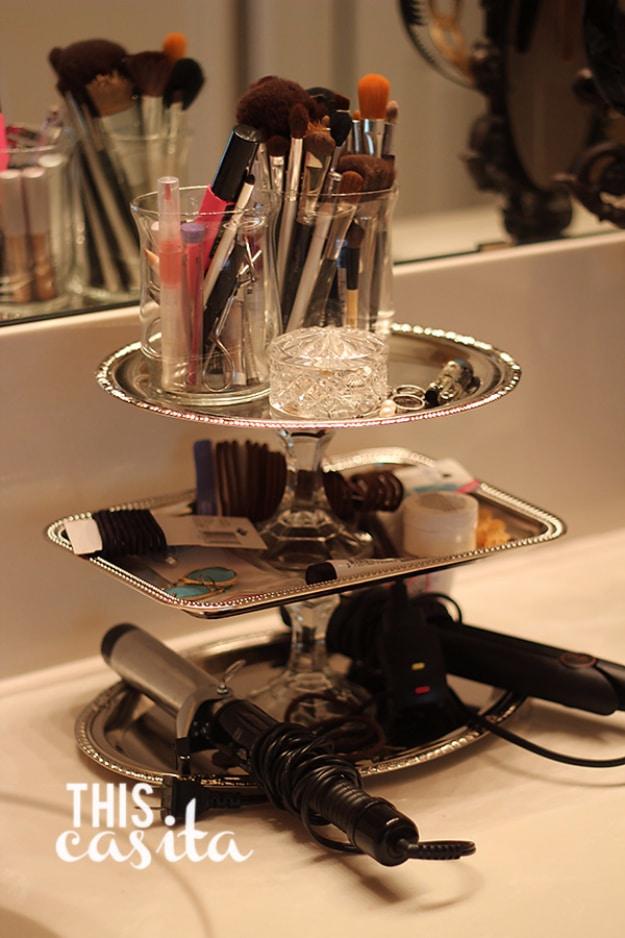 Three tier silver tray stand on a bathroom counter holding makeup brushes, hair ties, a curling iron and a flat iron