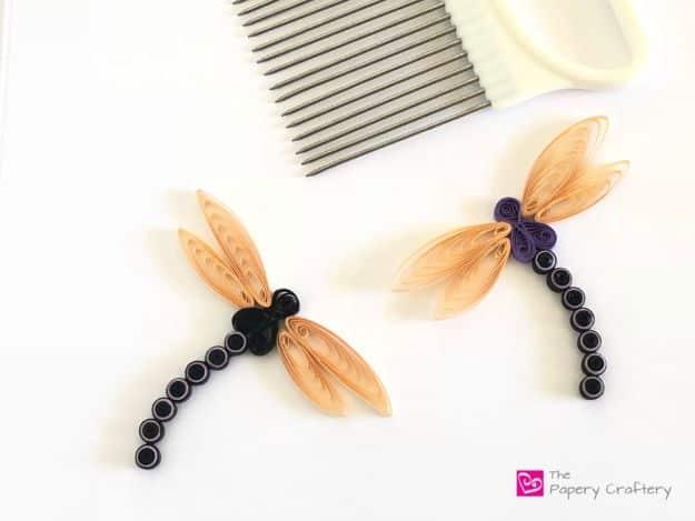 Two quilled paper dragonflies, one black bodied and one purple, with peach teardrop wings beside a quilling comb