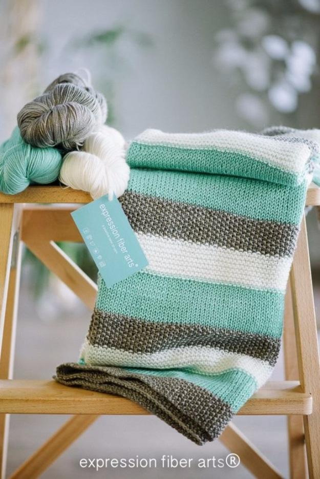 Mint, grey and white striped knitted baby blanket folded over a wooden rack with three skeins of yarn behind it
