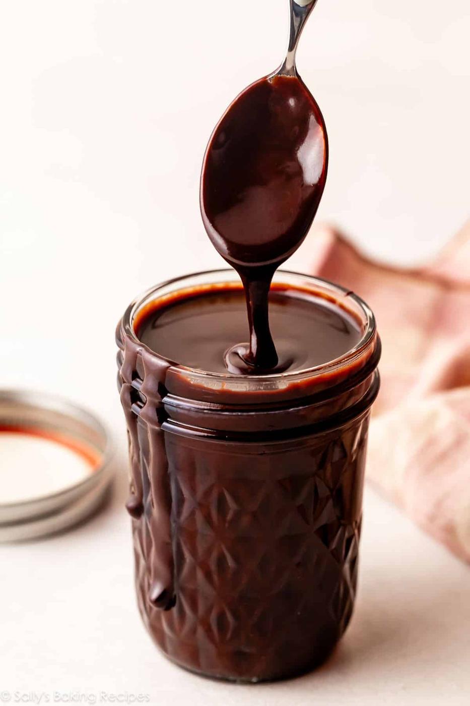 Small glass jar filled with thick glossy hot fudge sauce with a spoon lifting a thick ribbon of it out