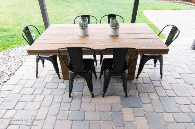 DIY outdoor dining table in stained wood with six black metal chairs around it, standing on a paver patio
