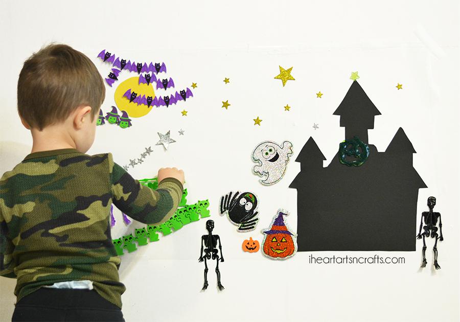 A young boy sticking paper bats, a ghost, skeletons and stars onto a contact paper haunted house scene on the wall