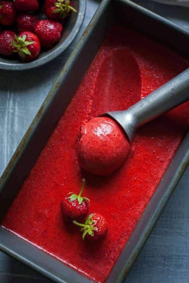 Deep red strawberry sorbet in a metal loaf pan being scooped, with a bowl of whole berries beside it