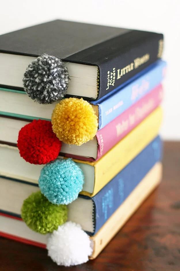 Yarn ball bookmark, colorful yarn pom poms dangling on strands from a stack of hardback books on wood