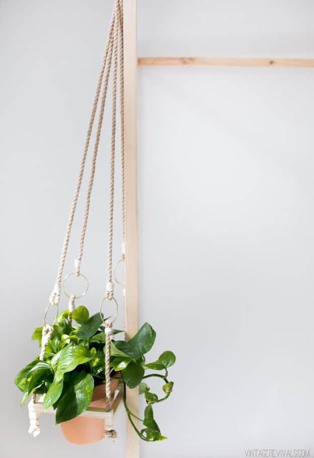 Square wooden hanging planter on rope, holding a terra cotta pot of trailing green plant beside a wall