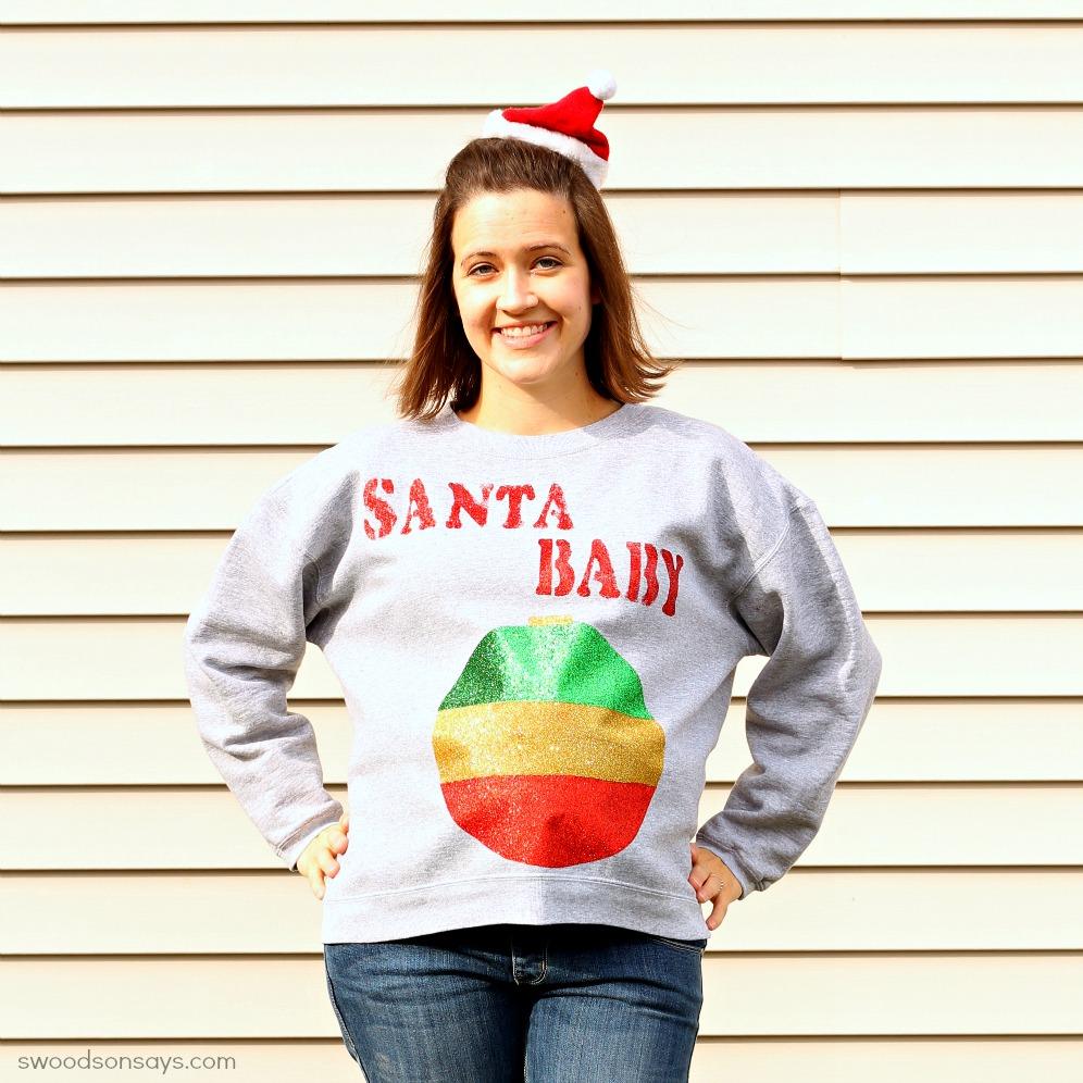 A pregnant woman in a santa hat wearing a gray sweatshirt with Santa Baby in red glitter above a big glitter ornament