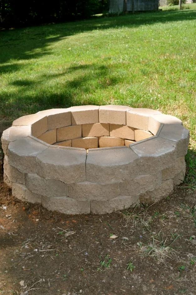 Back yard fire pit built as a ring of tan and gray landscape blocks on bare dirt at the edge of a grass yard