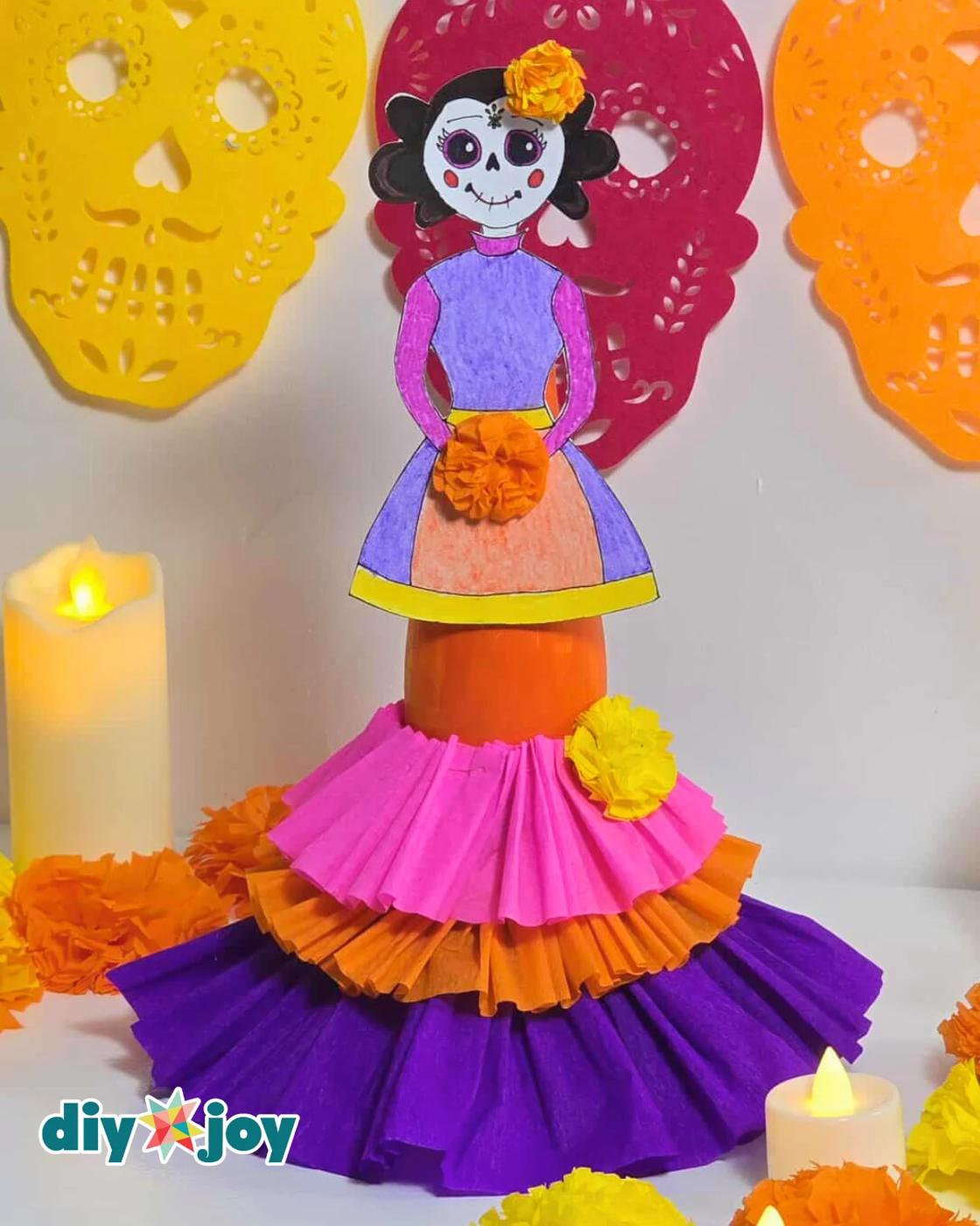 Day of the Dead Decorations - tight vertical crop of the finished La Catrina against a yellow, red and orange papel picado skull banner, with one lit candle and marigolds at the ed