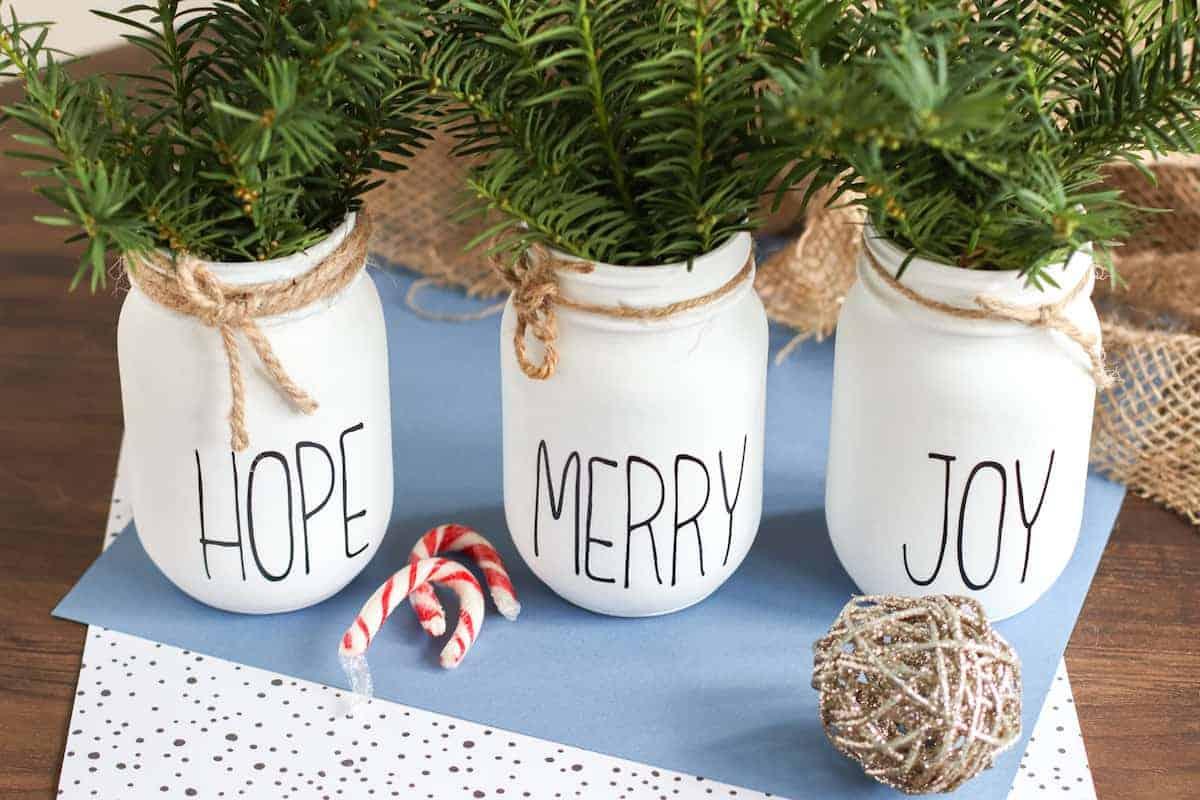 Three white painted mason jars lettered HOPE, MERRY and JOY in black, tied with twine and filled with fresh evergreen sprigs