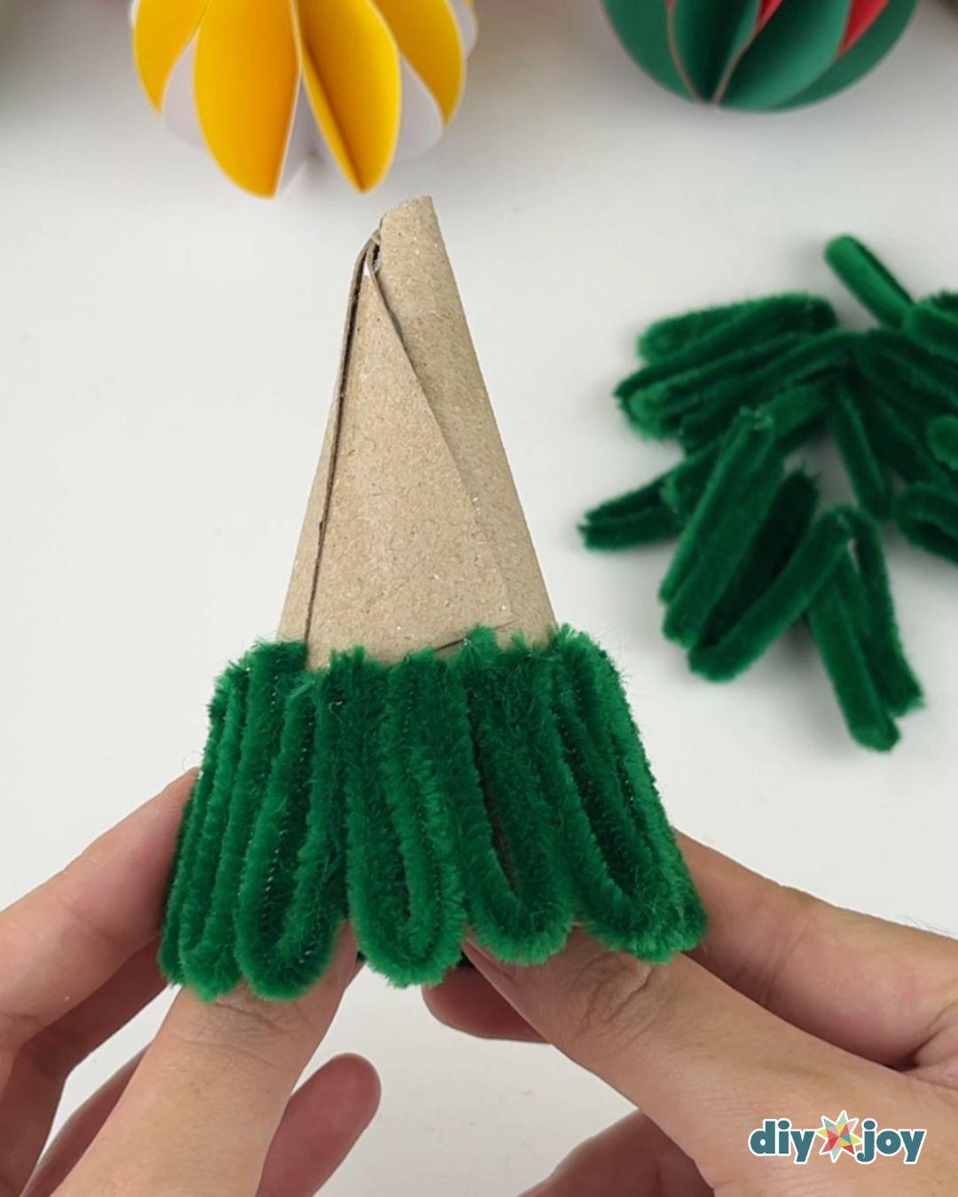 Pipe Cleaner Crafts - two hands hold a cardboard cone with the first row of folded green pipe cleaner pieces glued around the bottom rim