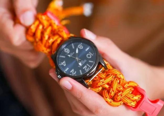 Hands weaving a bright orange paracord watch band onto a navy faced field watch with a pink plastic buckle