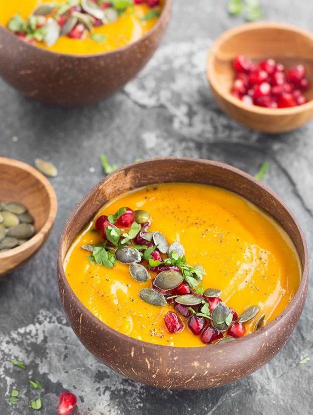 Wooden bowl of bright orange pumpkin apple soup topped with pomegranate seeds, pepitas and fresh herbs