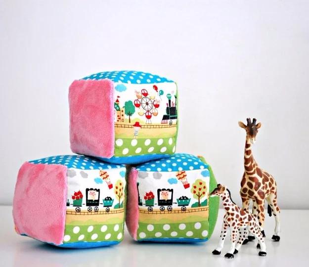 Three soft fabric rattle blocks with pink minky sides and a fairground print band, beside two toy giraffes