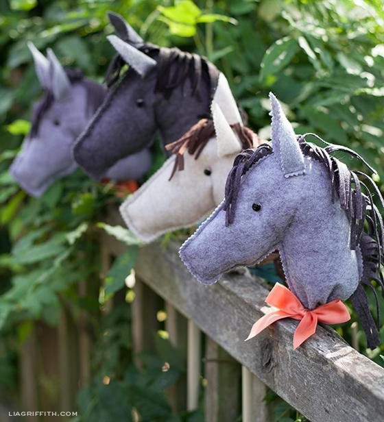 Three wool felt stick horse heads in gray, charcoal and oatmeal with fringed manes and an orange ribbon bow on a fence