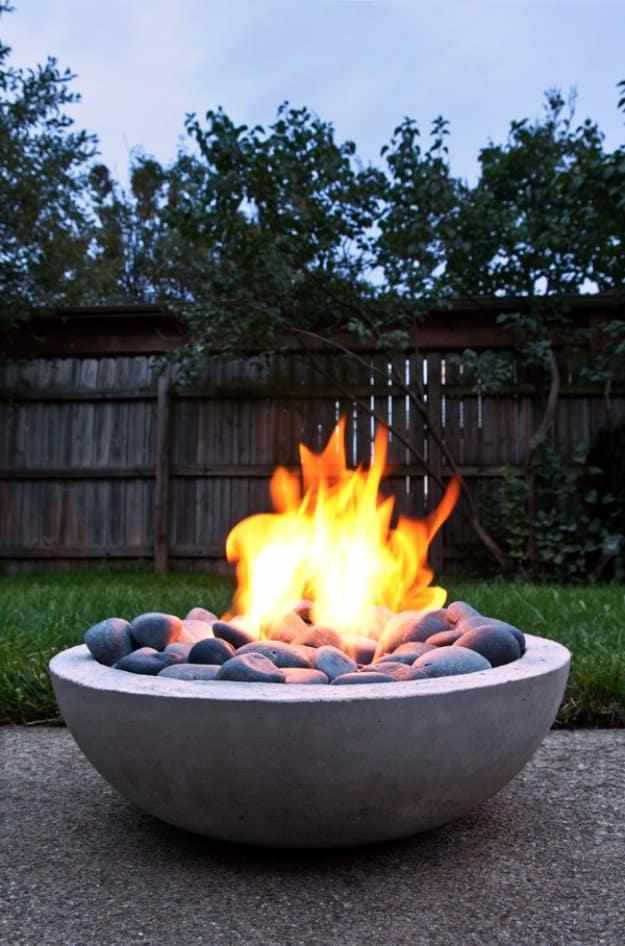 DIY concrete firepit, a round gray cast cement bowl full of black stones with flames burning on a patio at dusk