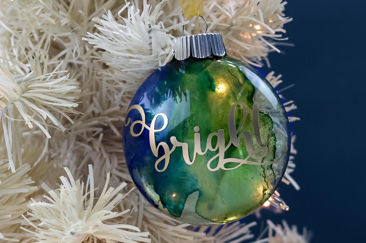 Clear ornament filled with blue and green marbled alcohol ink and a gold script word, hung on a white tree