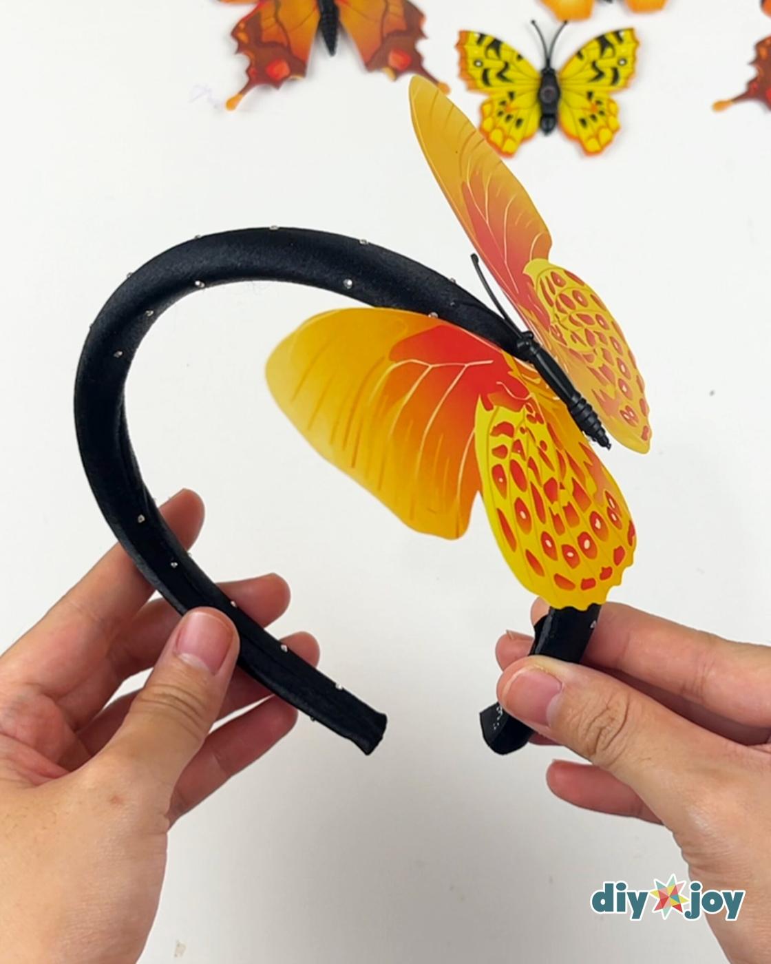 How to Make a Butterfly Headband - two hands gluing a fake butterfly onto a black headband with loose butterflies nearby