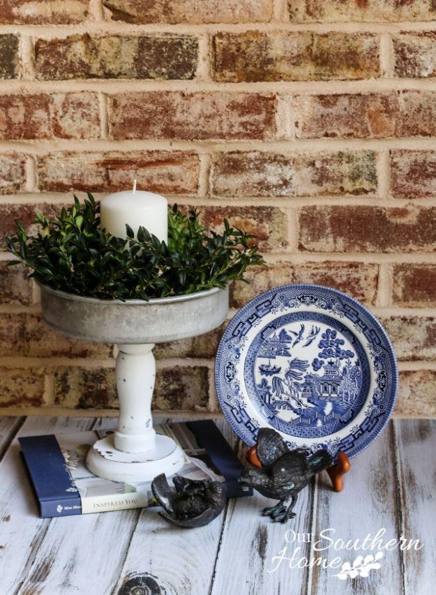 White painted cake pan and candlestick pedestal holding a candle and greenery against an exposed brick wall
