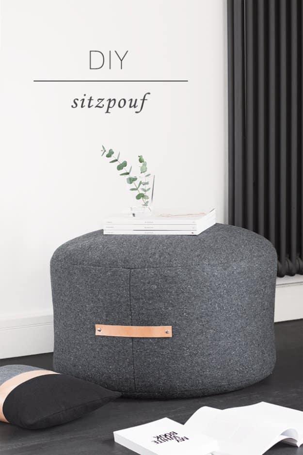 Low round grey wool pouf with a tan leather carry handle riveted to the side, on a dark floor by a white wall