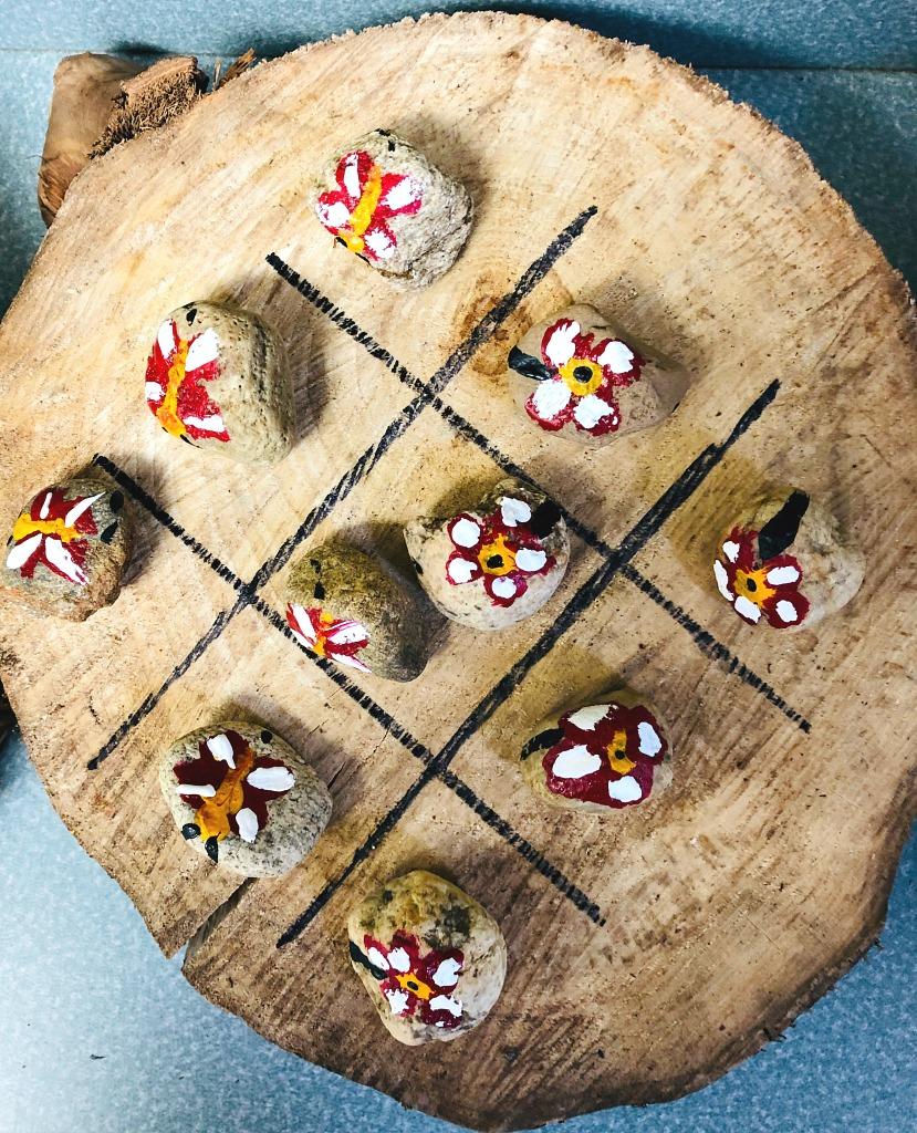 A tic tac toe game on a large wood slice with ten small rocks painted as red and white flowers and ladybugs