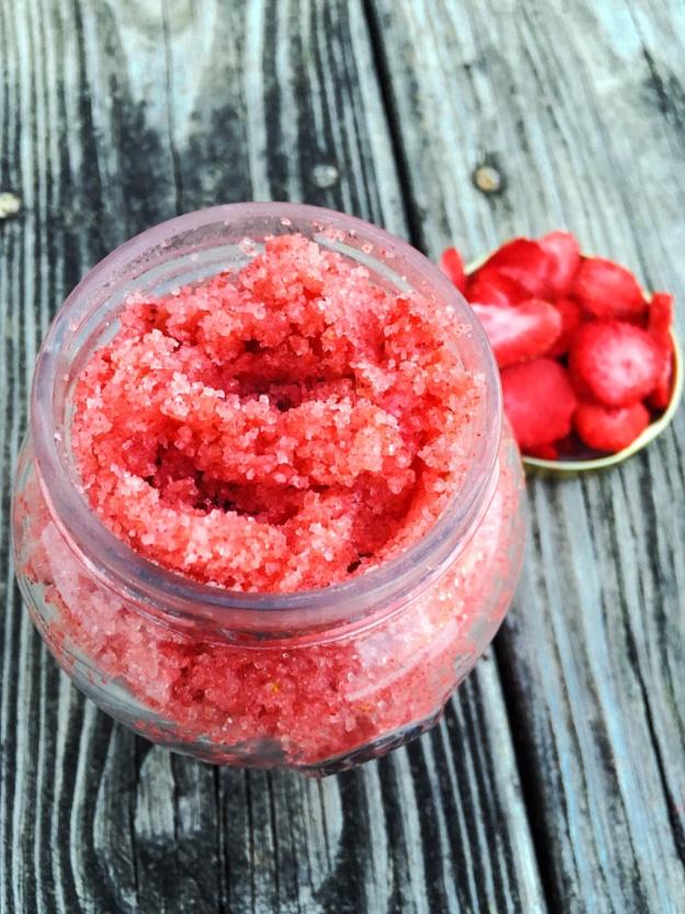 DIY strawberry sugar scrub, a bright red sugar scrub in an open jar with dried strawberries on teal wood
