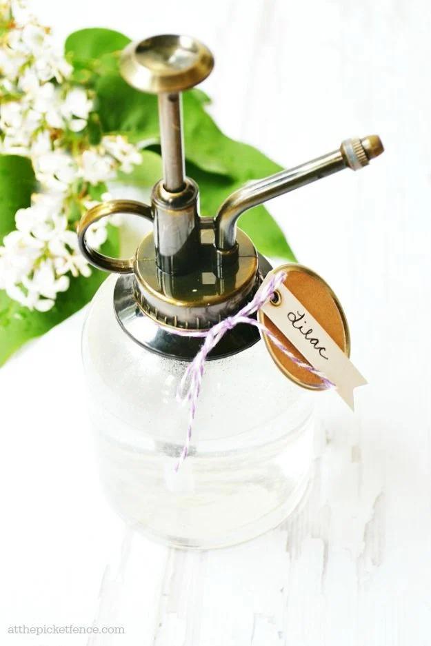 Glass misting bottle of homemade lilac room spray with a paper tag, sitting in front of white lilac blooms