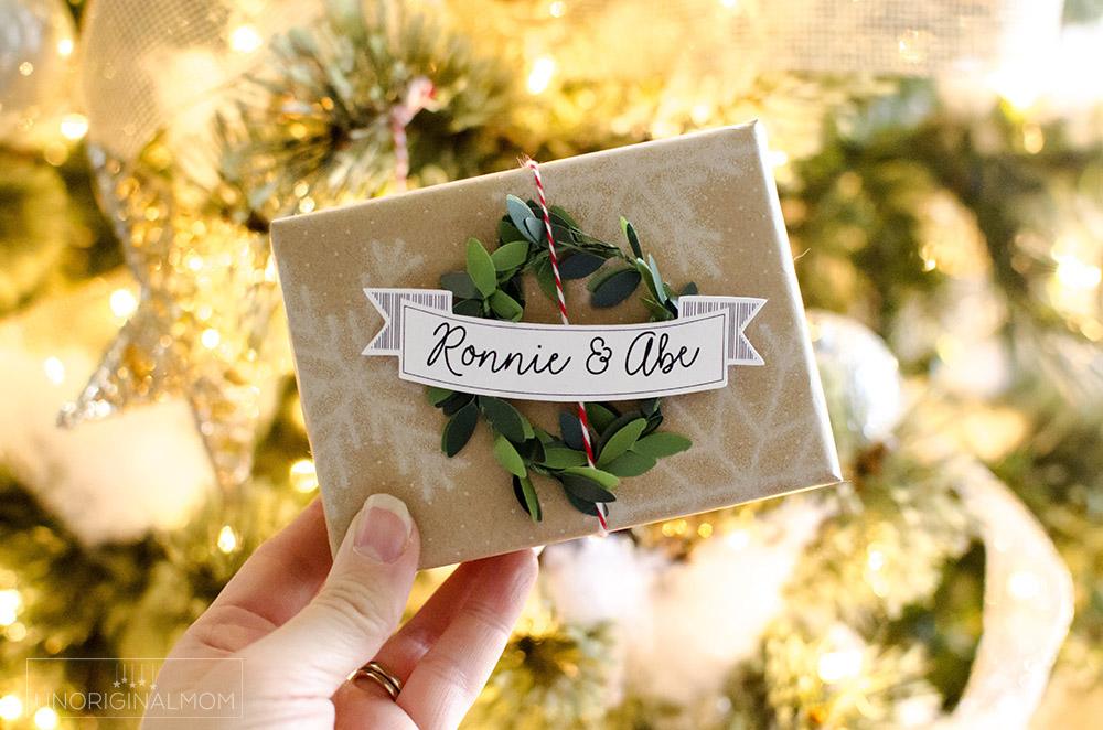 A hand holds a kraft parcel with a tiny green wreath and a banner name tag in front of a lit Christmas tree