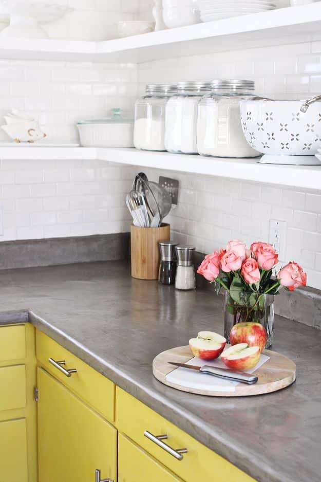 Grey concrete countertop over yellow lower cabinets with white subway tile, open shelves and pink roses