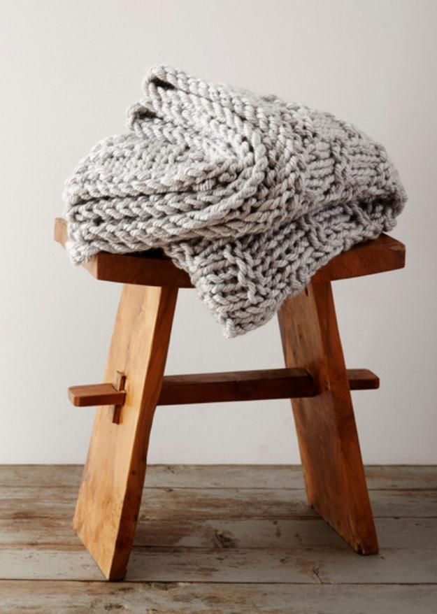 Thick pale grey chunky knit blanket folded over a small wooden stool on a bare board floor against a white wall