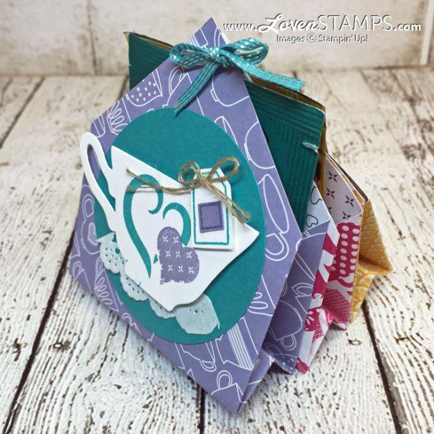 Folded lilac and teal patterned paper pocket holder with a teacup die cut, ribbon bow and twine, on white weathered wood