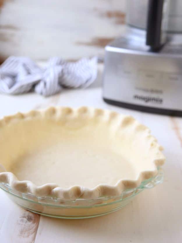Unbaked crimped pie crust in a glass pie plate on a white table with a food processor standing behind it
