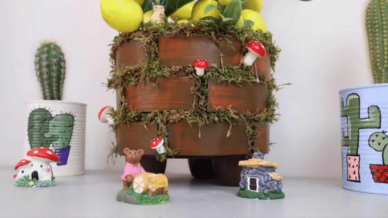 Tiered faux brick planter made from painted foam plates with moss and small red toadstools, beside cactus pots
