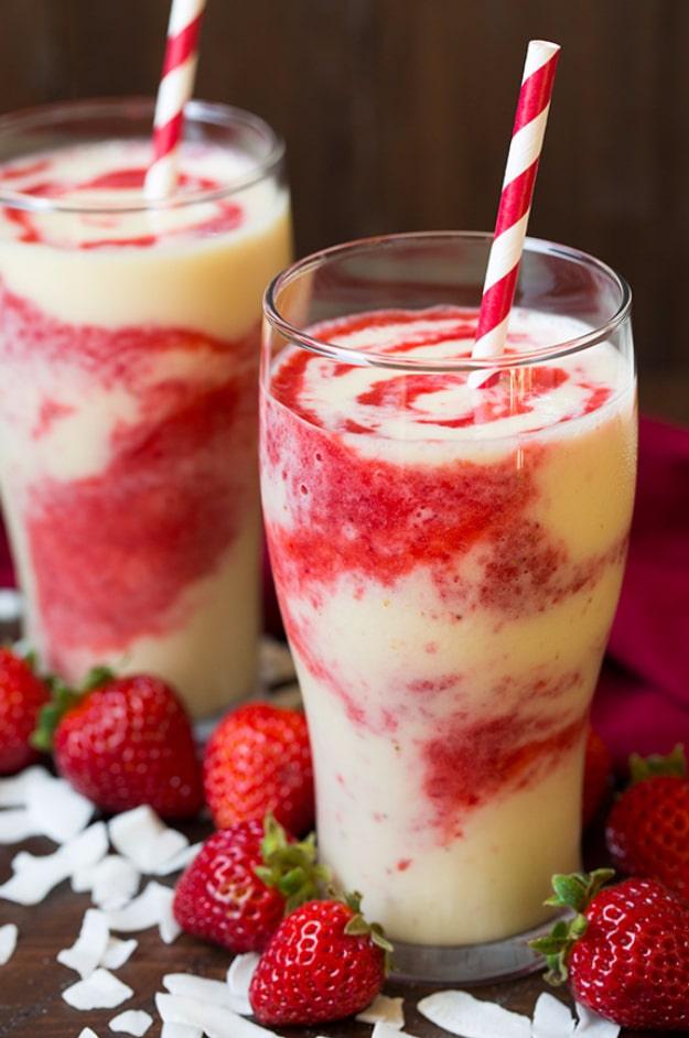 Two tall glasses of creamy coconut smoothie swirled with red strawberry puree, paper straws, strawberries and coconut flakes