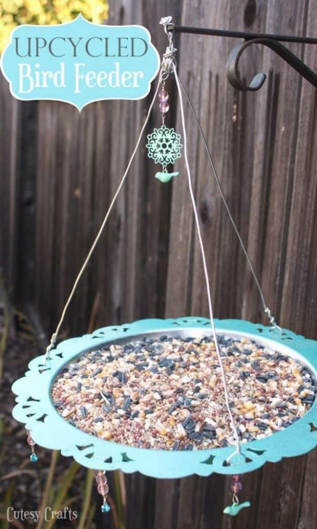 Upcycled silver platter bird feeder painted teal, strung with beads and bird charms on wire, full of seed by a fence