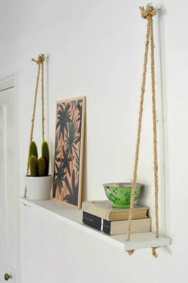 Wooden shelf hanging from knotted white rope on a wall, holding framed art, a green bowl and two bottles
