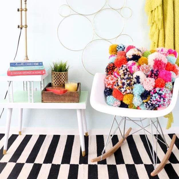 Throw pillow completely covered in bright yarn pom poms sitting on a white chair beside a striped rug