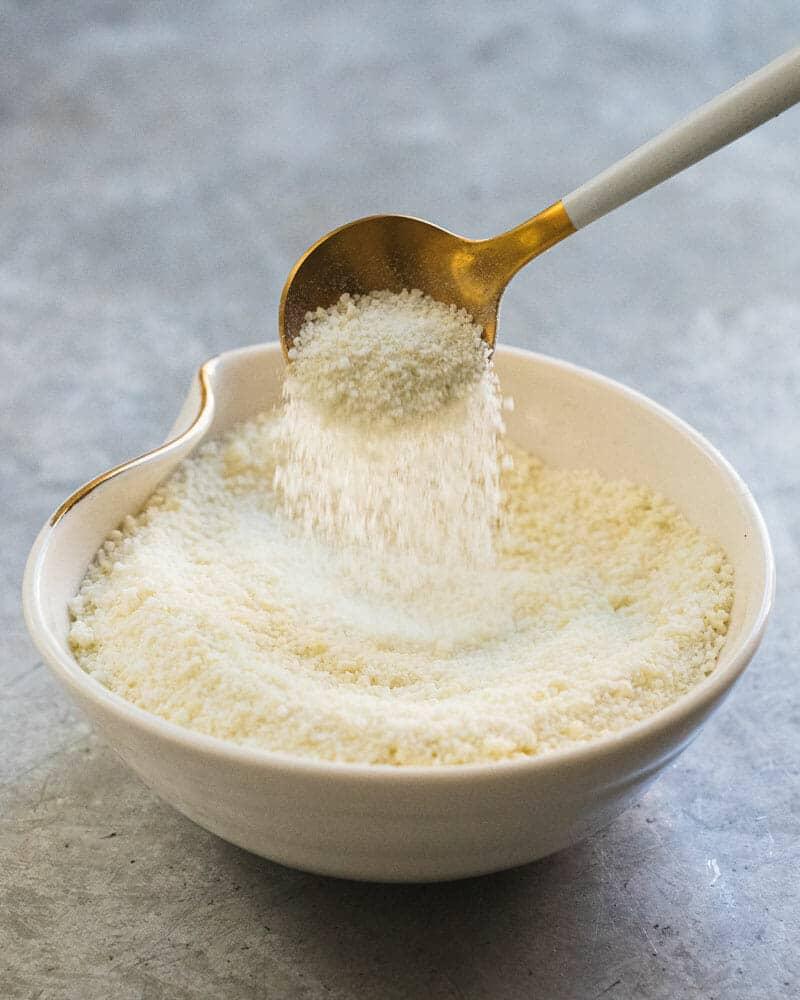 Gold spoon pouring fine white homemade garlic salt into a small white ceramic bowl on a gray stone counter