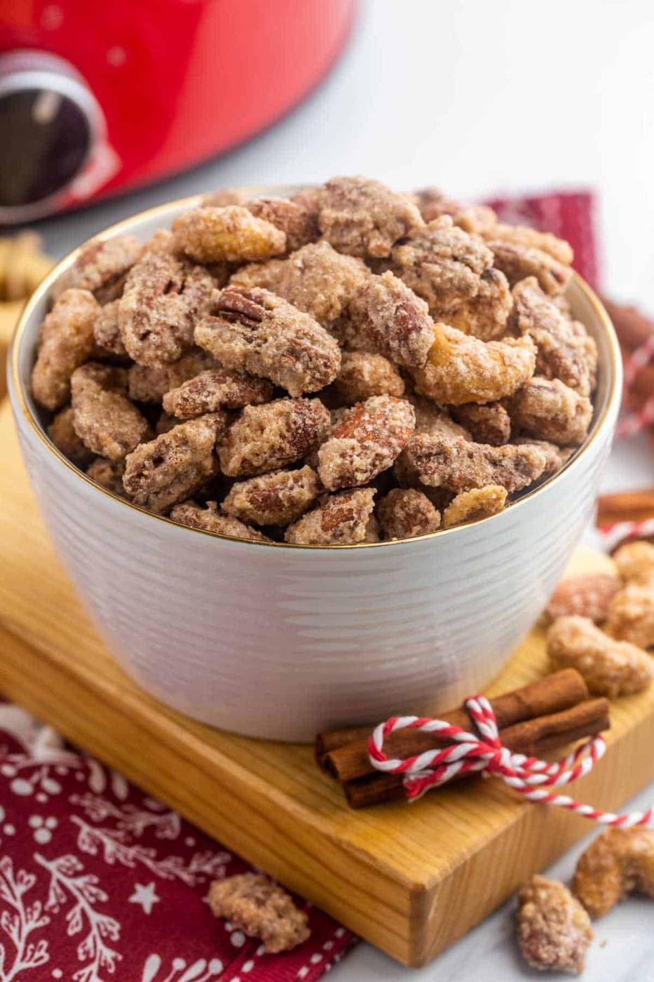 Cinnamon-sugar candied nuts in a bowl on red holiday textiles beside a slow cooker and cinnamon sticks