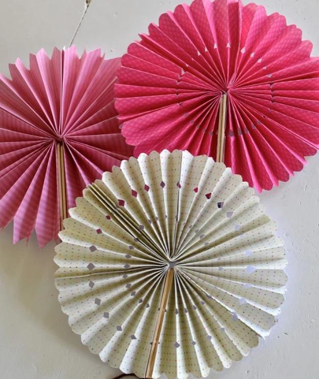 Paper pinwheel fans in hot pink, pale pink and cream with gold dots, mounted on sticks against a white wall
