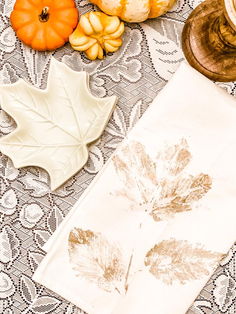 White tea towel printed with gold leaf shapes laid on a lace cloth beside small pumpkins and dried leaves