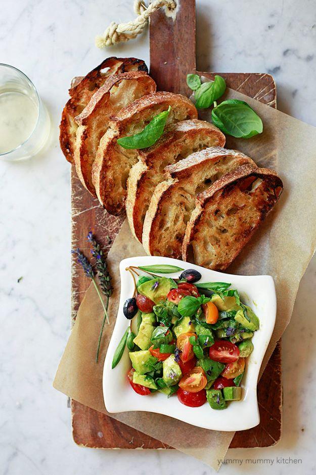 Avocado bruschetta with toasted baguette slices on a wooden board beside a white bowl of diced avocado and tomato