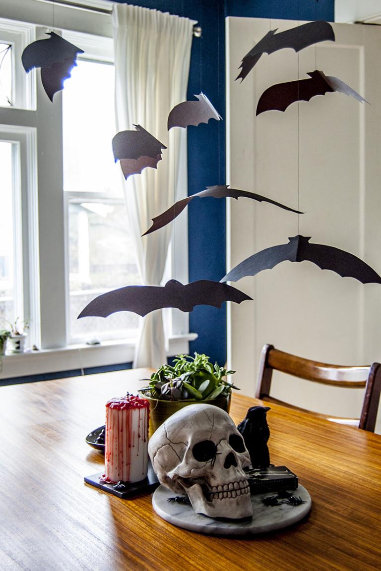 Black paper bats suspended on thread over a dining table with a skull and candle decorations beneath the hanging flock
