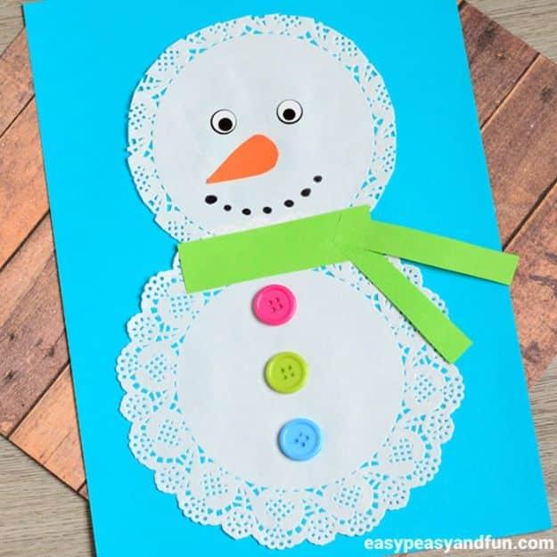 Doily snowman on bright blue paper with a green paper scarf, colored buttons, orange nose and googly eyes