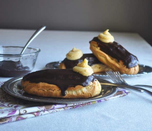 Chocolate eclairs glazed dark and finished with piped cream rosettes on a pewter plate with forks alongside