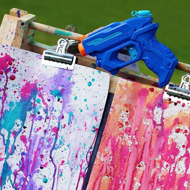 Blue squirt gun resting on a wooden easel above two sheets of paper splattered in pink, turquoise and purple paint