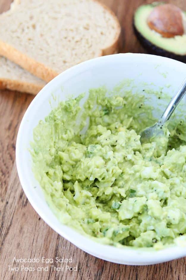 Avocado egg salad in a white bowl with a spoon, pale green and chunky, bread and half an avocado beside