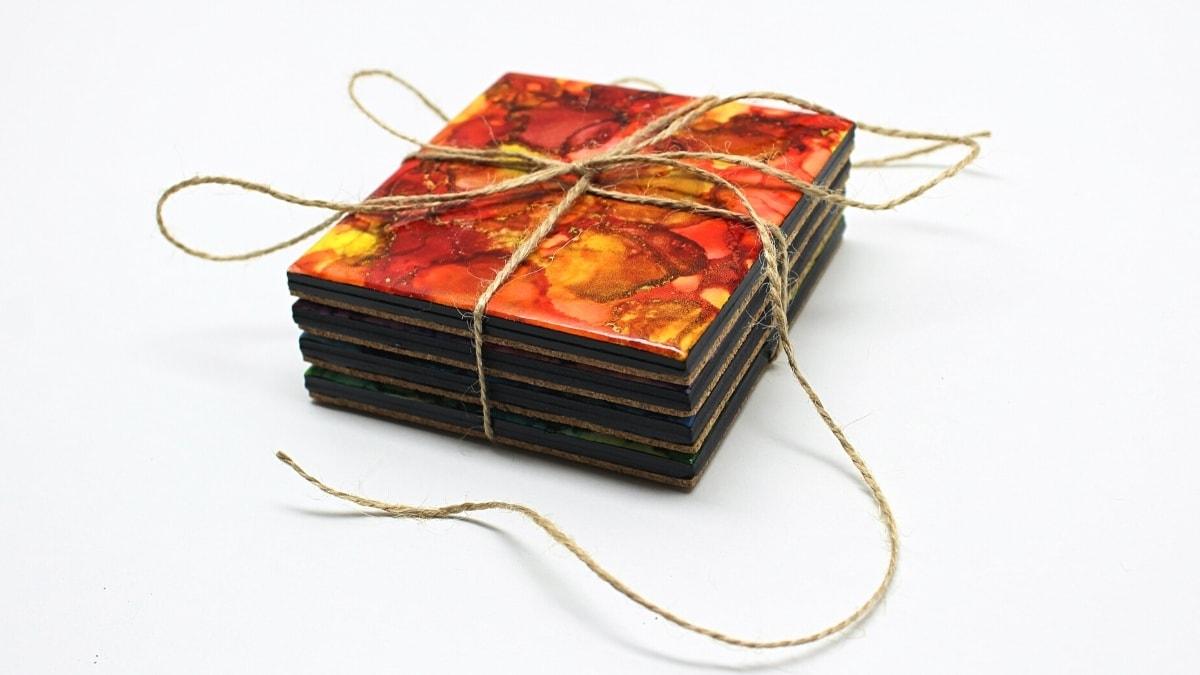 A stack of four ceramic tile coasters marbled in red, orange and gold alcohol ink with black edges, tied with jute twine