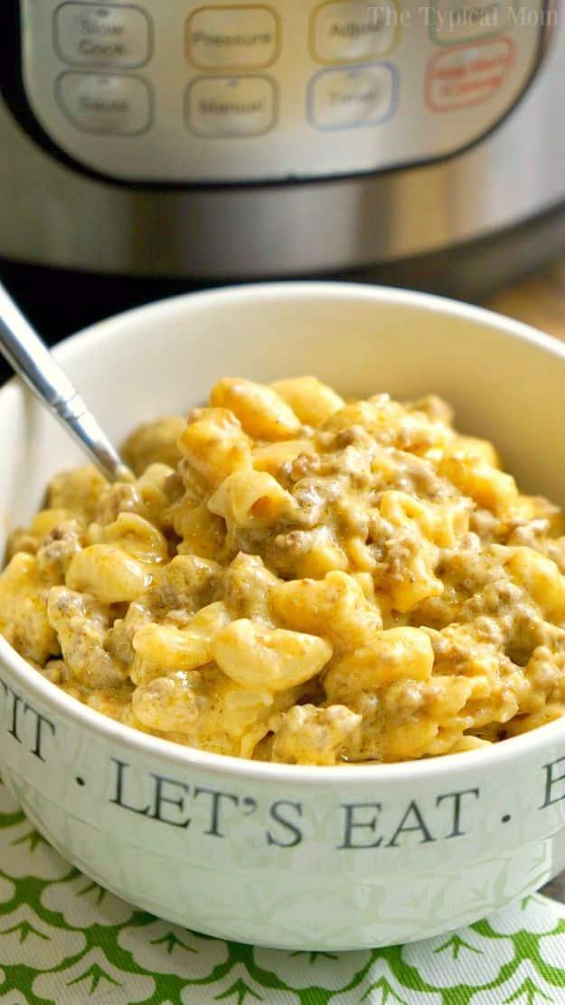 White bowl reading Lets Eat filled with cheeseburger macaroni with a spoon, an instant pot behind on a green cloth