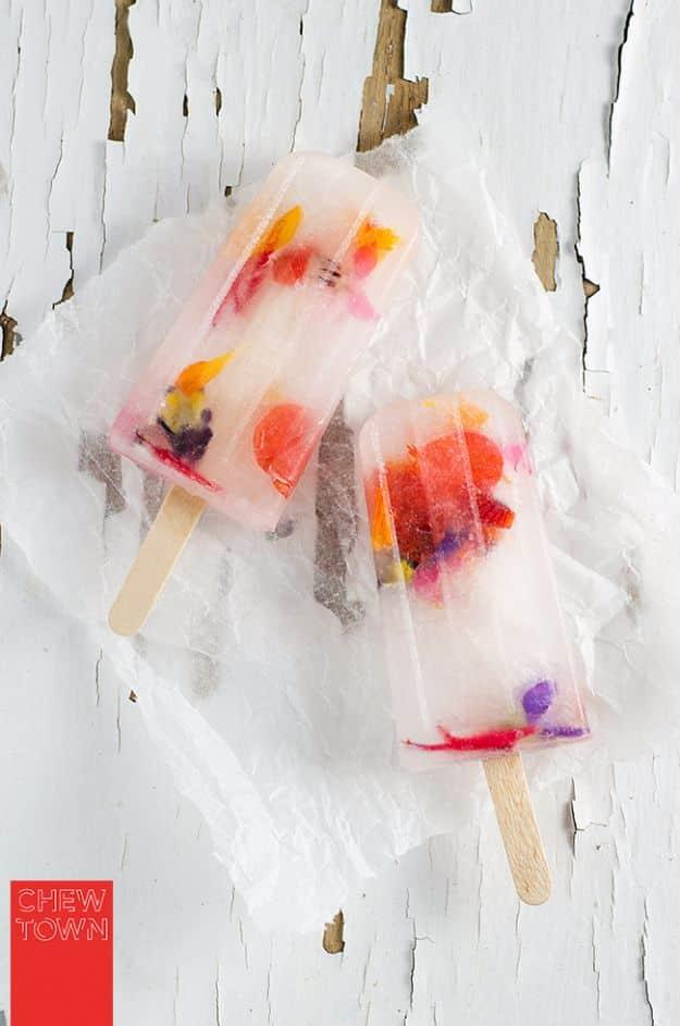Edible flower and elderflower popsicles, two clear ice pops set with coloured petals lying on crumpled white paper