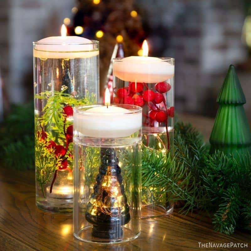 Three glass cylinders of water with floating candles on top and greenery, red berries and a lit mini tree submerged inside